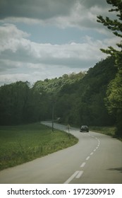 A single black car going down a curving road with tall green trees and a field of grass next to it