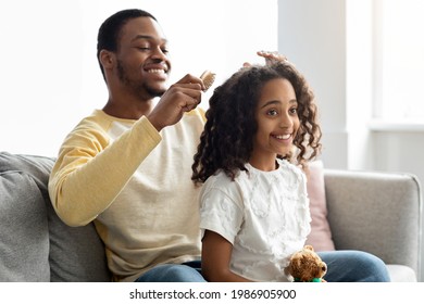 Single Black Father Combing Hair Of Happy Daughter While Sitting Behind Her On Couch At Home, Copy Space. Smiling African American Dad Brushing His Girl Kid Beautiful Curly Hair. Fatherhood Concept