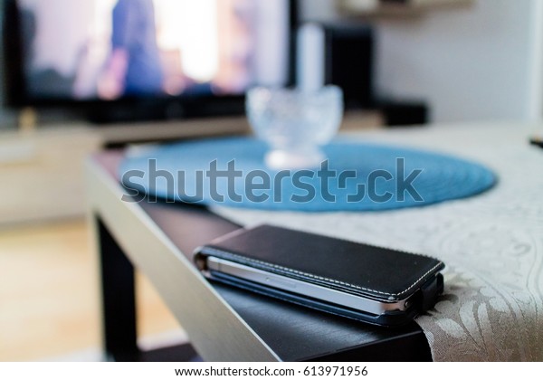 Single black leather\
case with cell phone on table next to dish over tablecloth in front\
of television