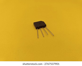 Single bridge rectifier diode with four metal terminals on yellow surface. High-quality electronic spare part for AC to DC power conversion and circuit board assembly.
