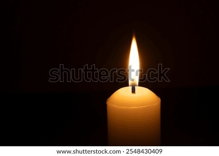 A single burning candle flame or light glowing on a big white candle is isolated on black or dark background on table in church for Christmas, funeral or memorial service