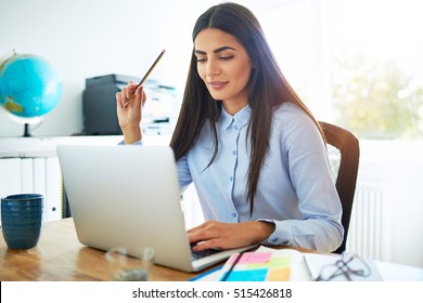 Single calm young Indian woman in blue blouse and long hair holding pencil in hand while seated at desk in front of laptop computer in bright room - Powered by Shutterstock