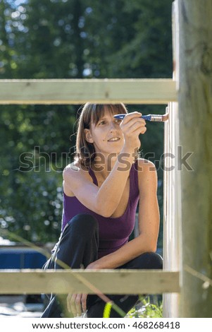 Single cheerful woman in purple sleeveless shirt framed by fence using brush to paint on wood.