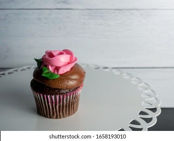 Single Chocolate Cupcake With Chocolate Frosting Decorated With A Pink Rose Made From Frosting Sitting On A White Decorative Platter Against A Gray Shiplap Background With Black Slate Surface.