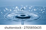 A single clear water drop is captured mid-fall, about to impact a calm blue water surface. Concentric ripples are expanding outwards from the impact point.