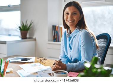 Single confident smiling woman in blue shirt with folded hands at desk in a bright, sunlight filled professional home office - Powered by Shutterstock