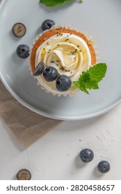 A single cupcake topped with piped white frosting, fresh blueberries, chia seeds, and a mint leaf sits on a gray plate under soft light.