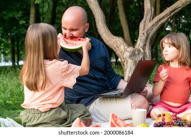 Single Dad Working Remotely On Laptop In Park. Raises Two Little Daughters. The Eldest Is Feeding Her Dad Ripe Red Watermelon.