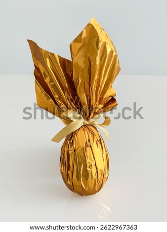 A single decorative Easter egg wrapped in glossy metallic gold foil and tied with a soft satin ribbon bow, isolated against a clean white background. The composition emphasizes minimalism and elegance, with reflective textures and subtle highlights enhancing the luxurious appearance of the wrapping material. The crinkled foil surface creates rich light reflections and depth, making the object visually striking and ideal for premium seasonal designs. This image is perfect for Easter-themed marketing campaigns, holiday promotions, packaging mockups, greeting cards, social media content, and festive branding materials. The neutral background provides ample copy space, allowing designers to easily integrate text or additional elements. The golden color palette conveys celebration, sophistication, and exclusivity, making it suitable for both traditional Easter contexts and modern luxury concepts. High detail and sharp focus ensure versatility across digital and print applications, including advertisements, banners, posters, and editorial layouts. The image also works well as a symbolic representation of surprise, gift-giving, and seasonal joy.