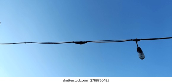 A single decorative outdoor string light bulb hanging from a black wire against a clear blue sky background.