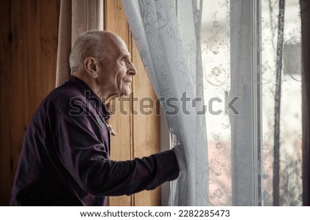 Single elderly man looking through window glass standing at home. Grandpa holding white curtain with hand and look out view from window.