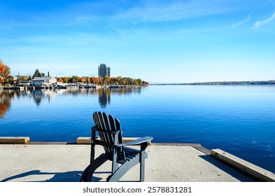 Single empty Adirondack chair on a cement pier overlooking a centered view of a shoreline marina surrounded by fall colors with blue rippling water in the foreground and a lightly clouded sky above. - Powered by Shutterstock - Get 15% off with code: PIKWIZARD15