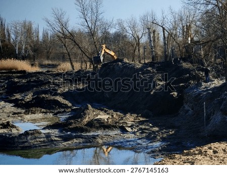 A single excavator digging dark soil and mud on a riverbank during ecological restoration works in early spring.