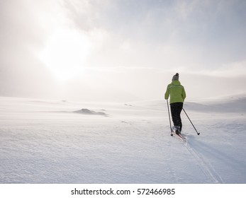 Single female cross-country skier off piste in the norwegian mountains with heavy clouds with the sun hiding at the edge passing some sun rays smoothly enlightening the flat terrain