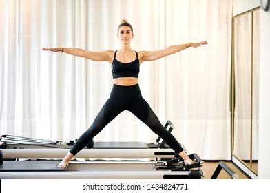Single fit woman doing yoga standing with arms and legs spread on reformer bed. White curtain in background.
