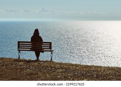 Single Girl In A Black Jacket And Hat Sitting On Bench At Cliff At Front Of Sea, Peaceful And Quiet Place For Thinking Alone, Loneliness And Loss Of Loved One Concept. Pacifying View Of Marine Horizon