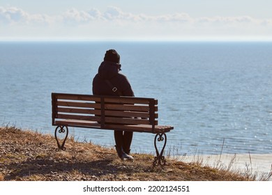 Single Girl In A Black Jacket And Hat Sitting On Bench At Cliff At Front Of Sea, Peaceful And Quiet Place For Thinking Alone, Loneliness And Loss Of Loved One Concept. Pacifying View Of Marine Horizon