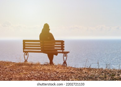 Single Girl In A Black Jacket And Hat Sitting On Bench At Cliff At Front Of Sea, Peaceful And Quiet Place For Thinking Alone, Loneliness And Loss Of Loved One Concept. Pacifying View Of Marine Horizon