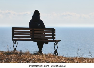 Single Girl In A Black Jacket And Hat Sitting On Bench At Cliff At Front Of Sea, Peaceful And Quiet Place For Thinking Alone, Loneliness And Loss Of Loved One Concept. Pacifying View Of Marine Horizon