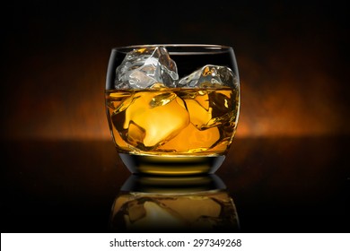 Single glass of whisky whiskey bourbon on ice with a reflective black surface and wooden background