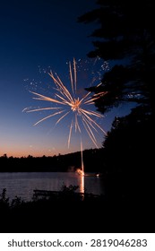 A single golden firework explodes above a still lake at dusk, framed by silhouetted pine trees, with the trail and burst reflected in the dark water.