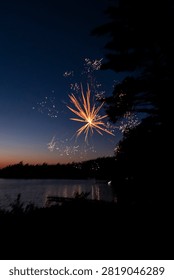 A single golden firework explodes above a calm lake at twilight with silhouetted pine trees framing the shore and light reflecting on the still water.