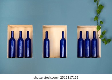 A single green ivy plant frames a row of  blue glass wine bottles arranged in an outcrop of a blue wall in a restaurant. 
