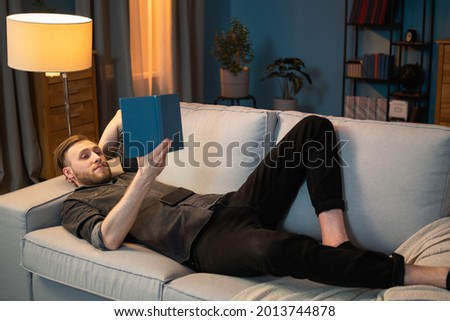 A single guy is hanging out after work in the home's cozy living room, lying on the couch with a blanket, hand propped under his head and holding a book, phone lying on his stomach
