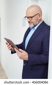 Single Handsome Middle Aged Bearded Man With Eyeglasses And Bald Head Looking Down At Tablet Computer While Leaning Against Wall With Copy Space