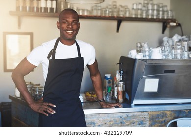 Single handsome young confident male coffee house owner with white shirt and black apron at counter with hand on hip