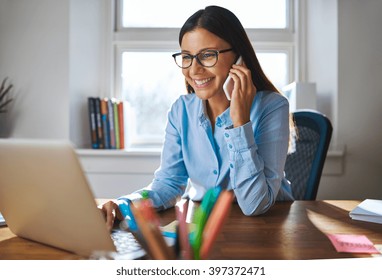 Single Happy Female Business Owner With Smile And Eyeglasses On Phone And Working On Laptop Computer At Desk With Bright Window In Background