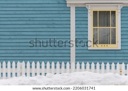 A single hung window in a blue exterior house wall of a vintage building. It has narrow v-grove clapboard on the wall. The window has yellow trim. There's a white picket fence in the foreground.