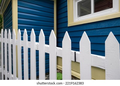 A Single Hung Window In A Dark Blue Exterior House Wall Of A Building. It Has Narrow V-grove Vinyl On The Wall. The Window Has A Yellow Sill. There's A White Picket Fence In The Foreground.