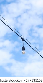 A single, illuminated Edison-style string light bulb hangs from a power cable against a backdrop of a bright blue sky with white, fluffy clouds. 