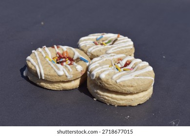 Single jam biscuit featuring white icing stripes and rainbow sprinkles. A perfect sugary snack or dessert isolated on a textured dark stone surface.