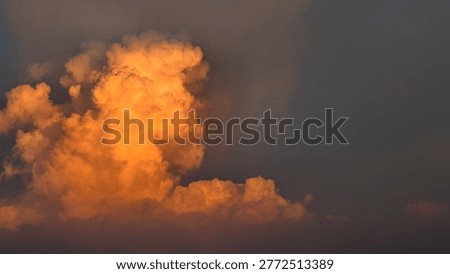 A single, large cumulus cloud is dramatically lit by the setting sun, casting a warm golden glow against a dark, moody sky.