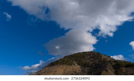 A single large white cloud floating above a forested mountain peak and a deep blue sky.