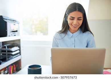 Single Laughing Young Indian Woman Beside Printer On Shelf And Wearing Long Hair While Seated In Front Of Laptop Computer In Bright Room