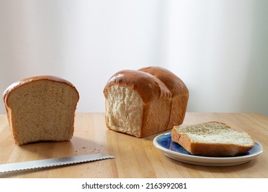 A single loaf of fresh white crusty bread on a wooden cutting board on a kitchen table. The warm crisp bun has melted butter over the crisp loaf buns. Side plates and a knife are next to the loaf.  - Shutterstock ID 2163992081