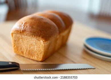 A Single Loaf Of Fresh White Crusty Bread On A Wooden Cutting Board On A Kitchen Table. The Warm Crisp Bun Has Melted Butter Over The Crisp Loaf Buns. Side Plates And A Knife Are Next To The Loaf. 