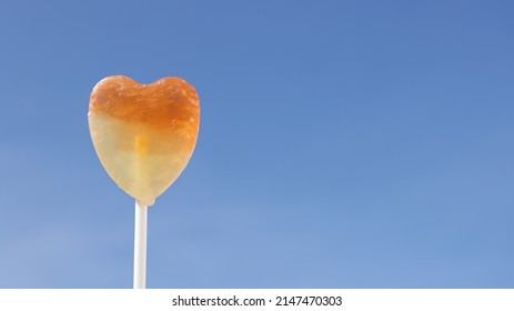 Single Lollipop In Heart Pattern Orange And Yellow, Blue Sky Background. Sweet Candy Hearts In Symbol Of Love, Copy Space