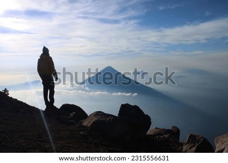 Single male hiker on top of Volcan de Acatenango with above the clouds view of Volcan de aqua in Guatemala. Person over clouds during rising morning fog and blue skies. Extreme altitude trekking.