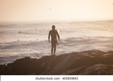 Single Man With Surf Standing At The Anchor Point In Morocco Waiting For A Wave During Sunset.