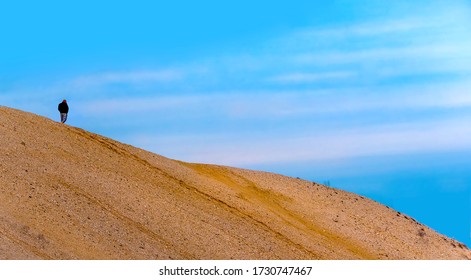 A single man is walking on the top of a huge sand dune with blue sky in the background.