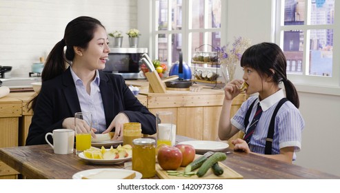 Single Mom And The Only Child Family At Breakfast Time In Wooden Kitchen Interior. Young Asian Businesswoman Manager Play Mother Role At Home. Parent Joy Talk To Kid While Little Girl Eating Sandwich