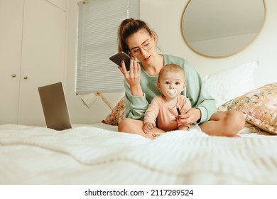 Single Mother Taking A Phone Call While Sitting On A Bed With Her Baby. Multitasking Mother Making Plans With Her Clients While Working From Home. New Mom Balancing Motherhood And Work.