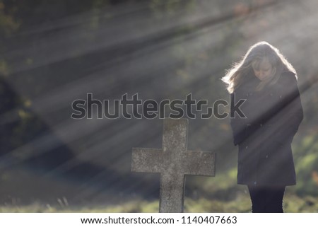 Single Mourner. Social distancing burial. Heavenly rays of sunshine behind grieving widow in cemetery. Mourning lady dressed in black contemplating life and death by the stone cross of a grave.
