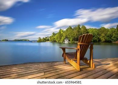 Single Muskoka chair sitting on a wood dock facing a lake. Across the calm water is a white cottage nestled among green trees. There is a boat dock on the water in front of the cottage.