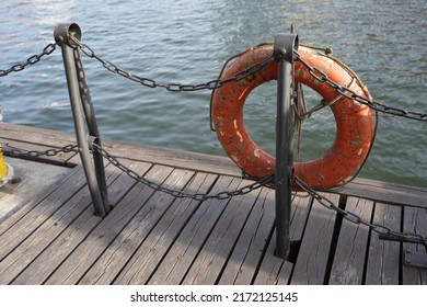 A single orange lifebuoy hanging on a metal fence with thick black metal chain on wooden desk with water in the background