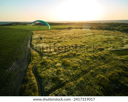 A single paraglider enjoys an exhilarating flight over expansive green fields under the clear blue sky.